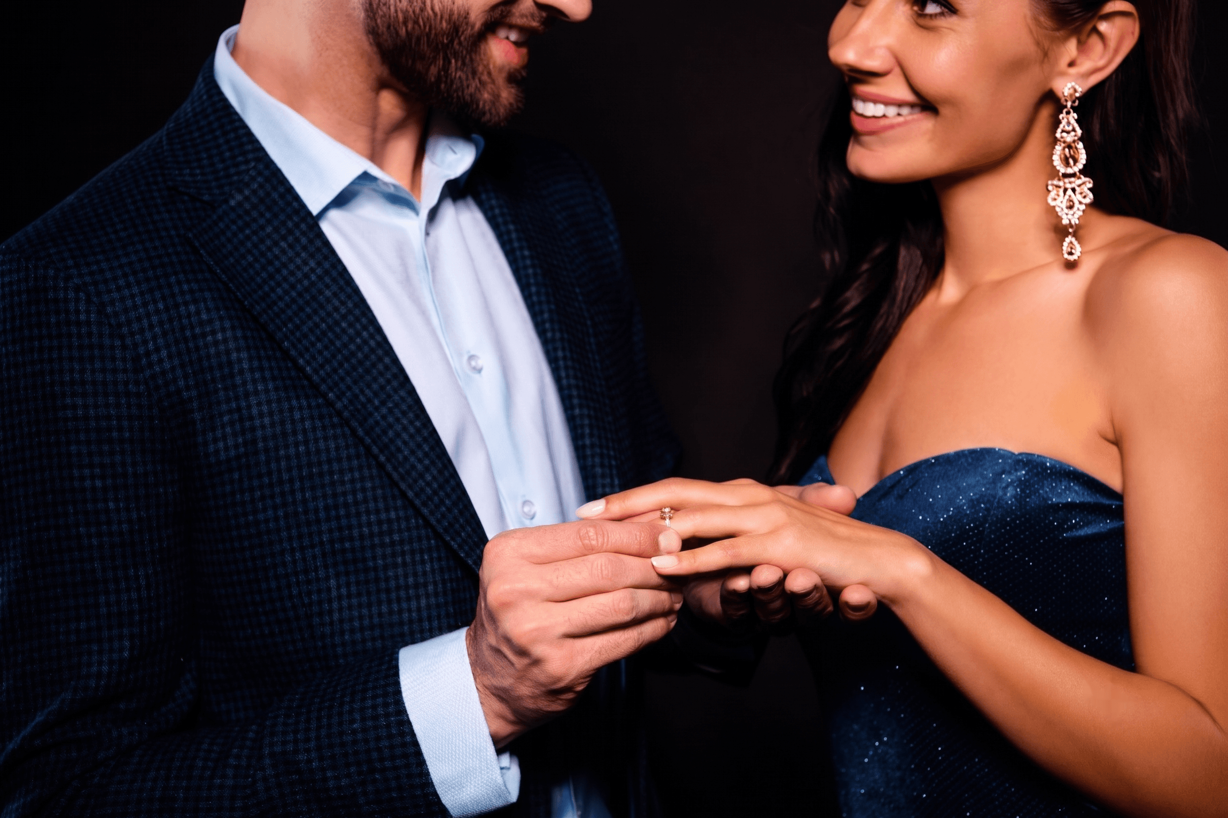 A man placing a gold engagement ring onto a woman's finger, both smiling at each other.