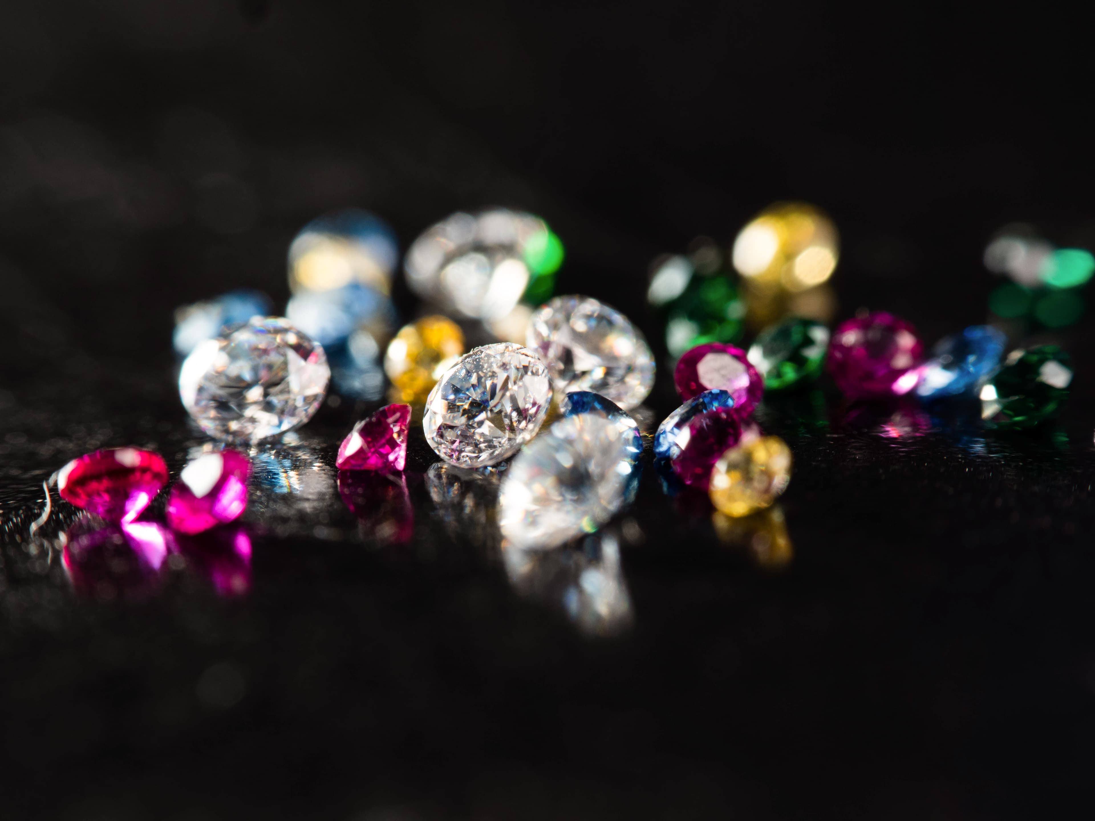 A curated selection of loose diamonds and coloured gemstones — white, ruby pink, sapphire blue, emerald green and golden yellow — scattered on a dark reflective surface